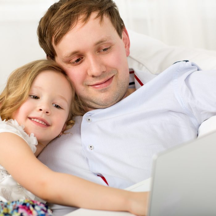 Little girl using laptop sitting on the sofa with father and leaning on his shoulder